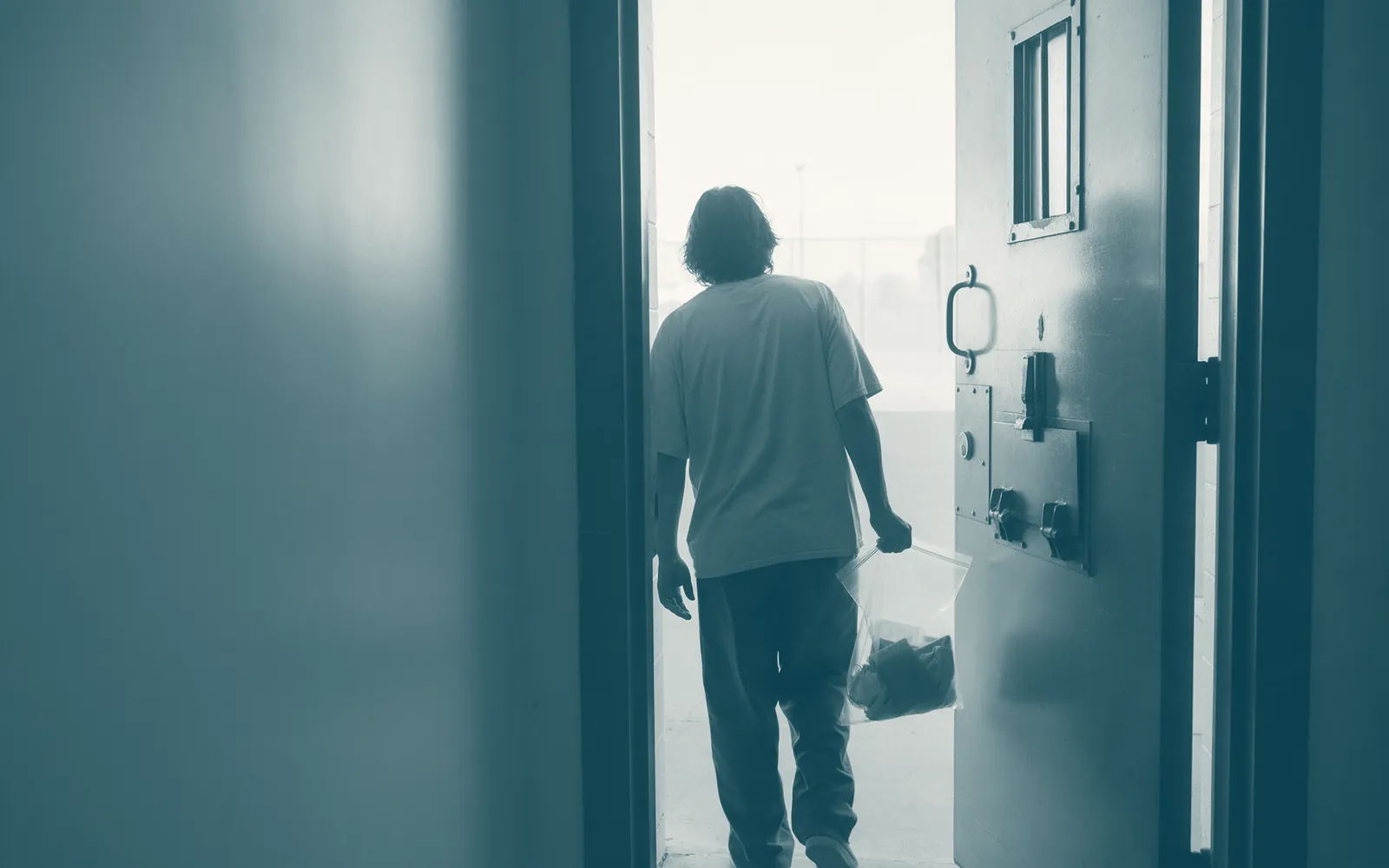A person stepping out of a facility door into morning light, carrying a property bag.